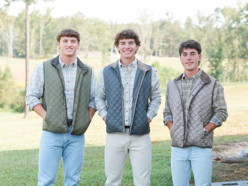3 men wearing vests and wrangler jeans smile at the camera in front of greenery