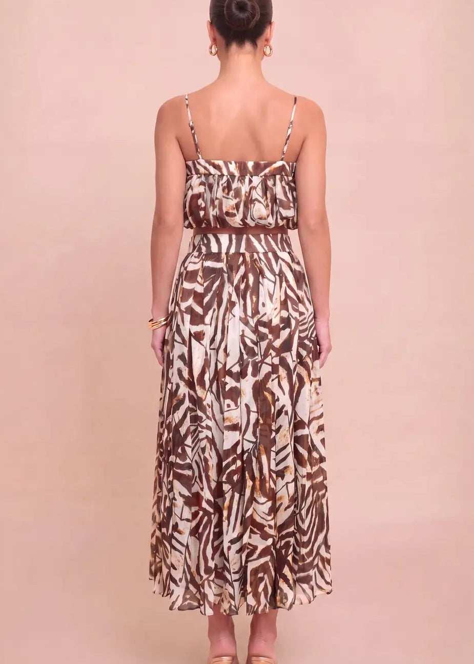 A woman viewed from the back wearing a sleeveless maxi dress with thin straps and an abstract brown and cream print, standing against a solid pale pink studio background.