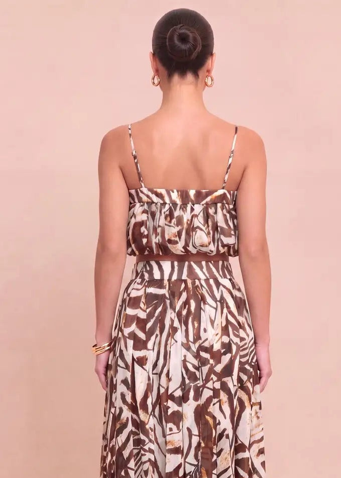 A woman viewed from the back wearing a sleeveless maxi dress with thin straps and an abstract brown and cream print, standing against a solid pale pink studio background.