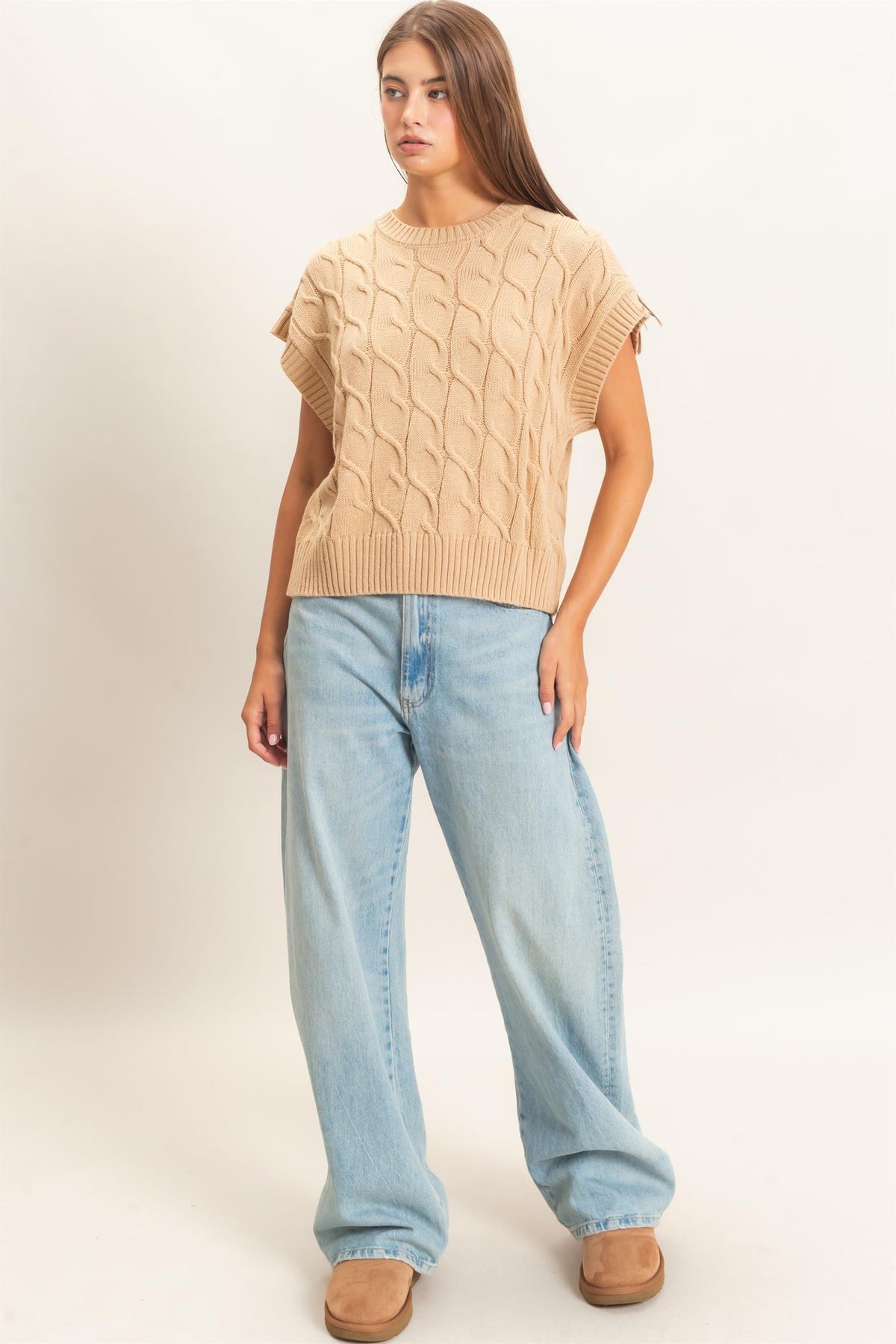 A woman modeling a beige cable-knit short-sleeve sweater paired with light-wash wide-leg jeans and brown shoes, standing against a plain off-white studio background.