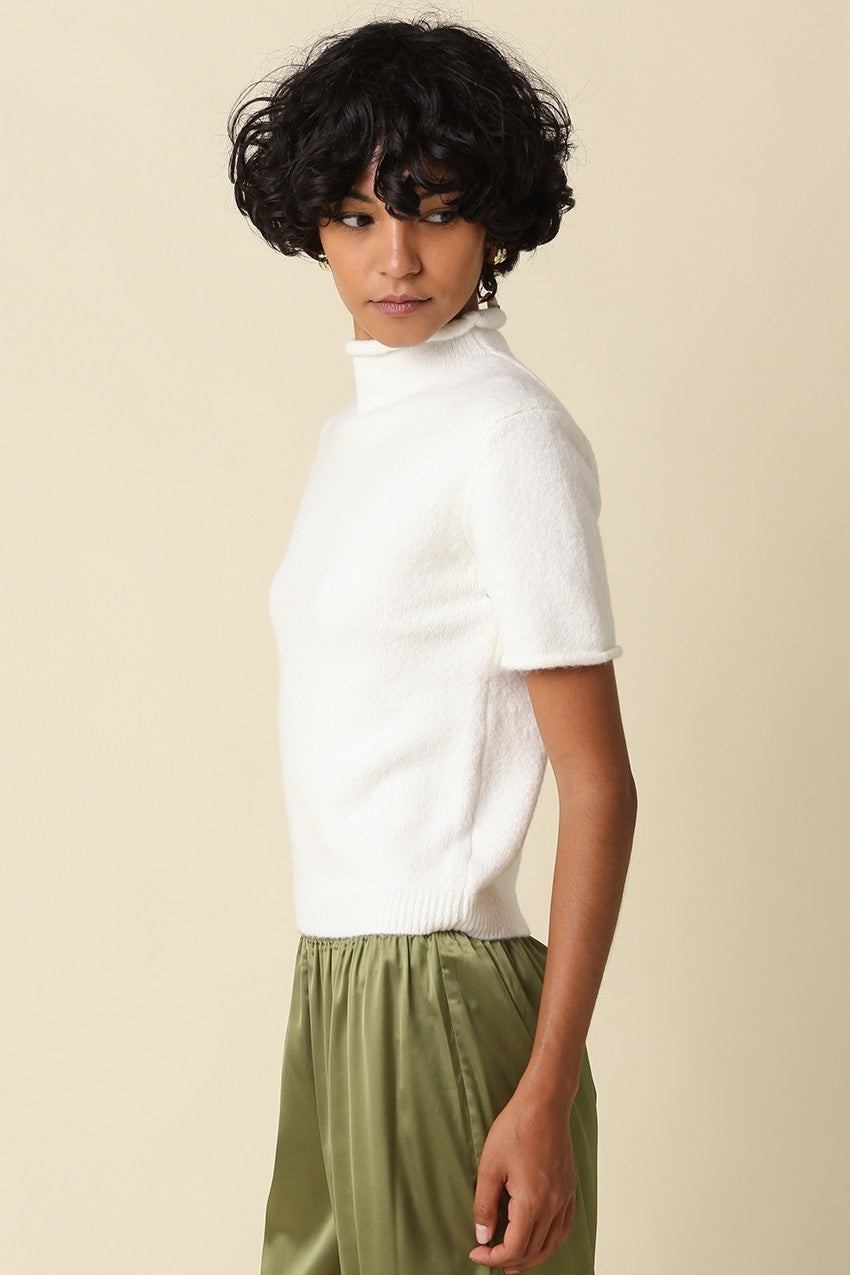A model wearing a white short-sleeve mock neck knit top and olive green satin trousers, photographed against a solid light beige background.