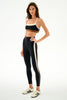 A model wearing a black sports bra with a white band and matching black leggings featuring a white and gold side stripe, paired with white sneakers, against a plain white studio background.