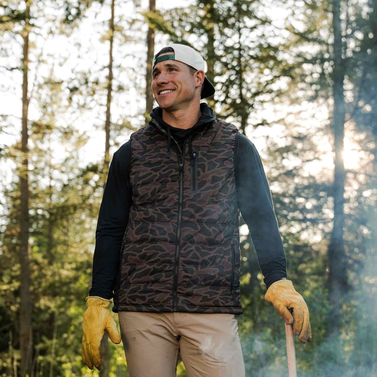 A man wearing a camouflage zip-up vest over a black long-sleeve shirt, khaki pants, and yellow gloves, standing outdoors in a forest with trees in the background.