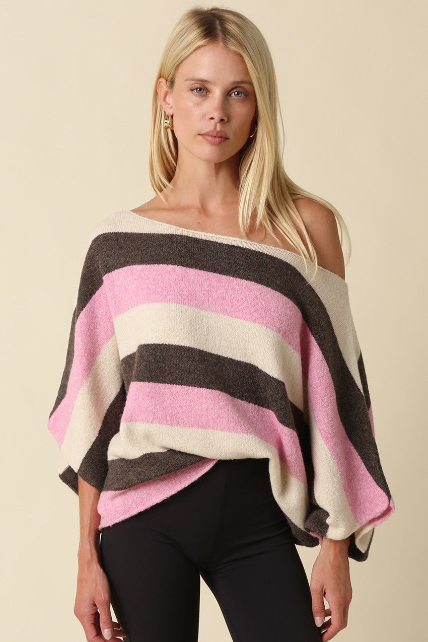 A woman modeling a knit poncho top with horizontal stripes in dark brown, light pink, and cream, paired with black pants against a solid beige background.