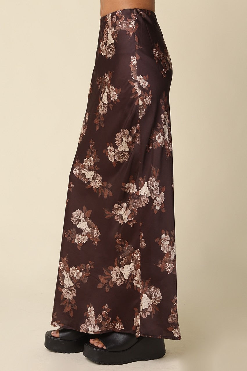 A person wearing a long, wide-leg skirt in dark brown satin with a beige and white floral print, paired with black platform sandals, shown from the waist down against a solid beige background.