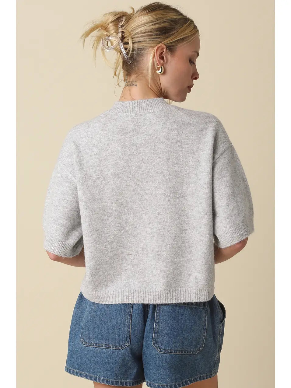 A woman shown from the back wearing a light gray short-sleeve knit sweater and blue denim shorts against a plain beige studio background.