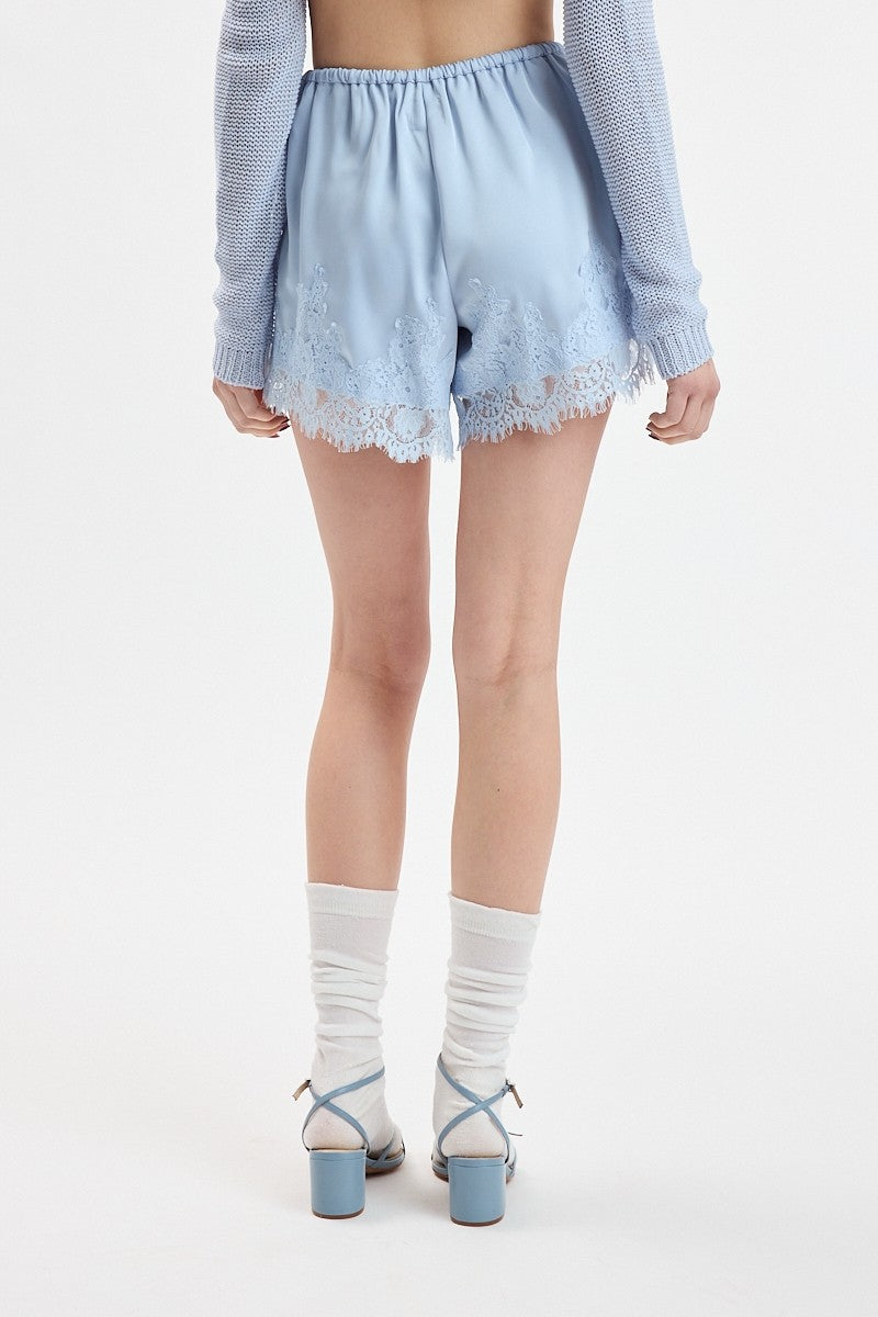 A model shown from the back wearing light blue satin shorts with a white lace hem, a light blue knit cardigan, white socks, and light blue block-heel sandals against a plain white studio background.
