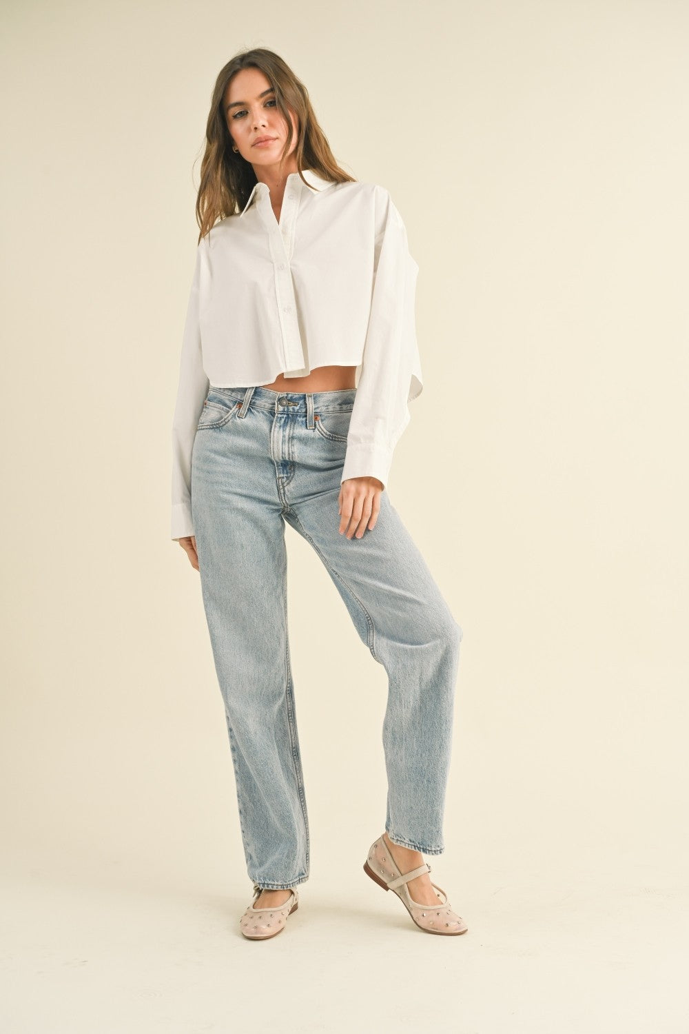 Woman standing on tan background wearing a cropped white button front shirt and jeans