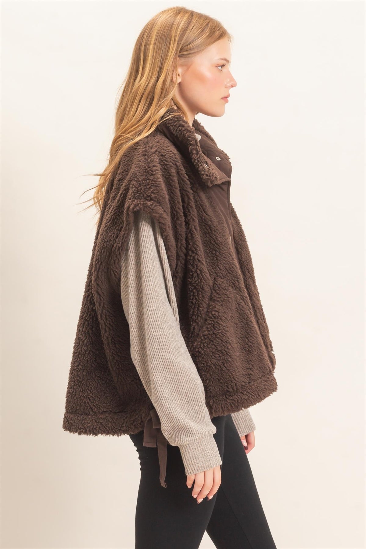 A woman shown in side profile wearing a brown sherpa fleece vest over a light beige ribbed knit sweater and black pants, photographed against a plain light background.