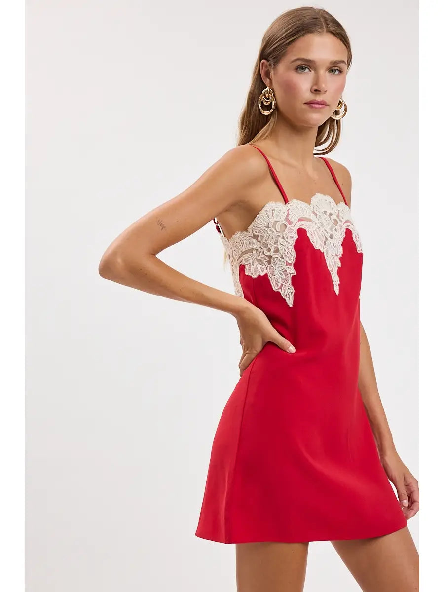 A woman modeling a red slip-style mini dress with thin straps and a white lace yoke, posed against a plain light gray studio background.