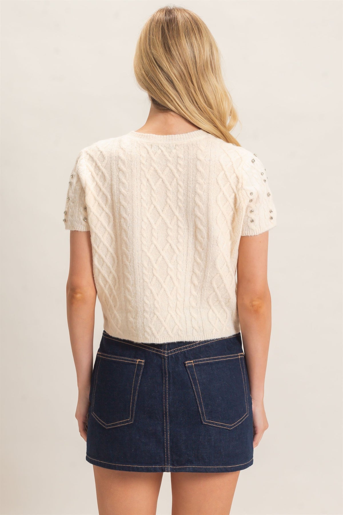 A woman shown from the back wearing a cream cable-knit short-sleeve top with lace sleeves and a dark blue denim mini skirt, against a plain light background.