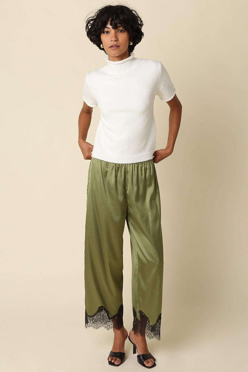 A model wearing a white short-sleeve mock neck top and olive green wide-leg pants with black lace trim at the hem, paired with black heeled sandals, against a solid beige background.