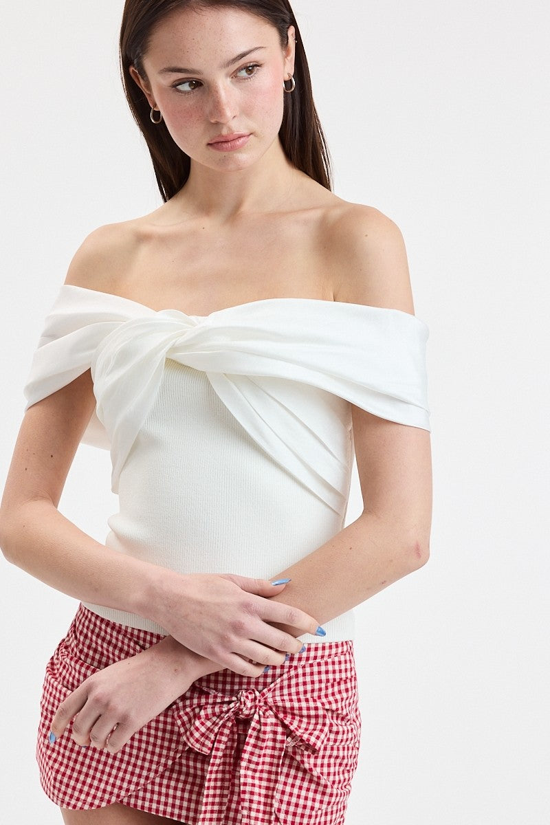 A woman wearing an off-the-shoulder white top with a large front knot detail and red-and-white gingham shorts against a plain light background.