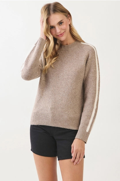 A woman modeling a beige knit sweater with white stripe details on the sleeves, paired with black shorts, against a plain white studio background.