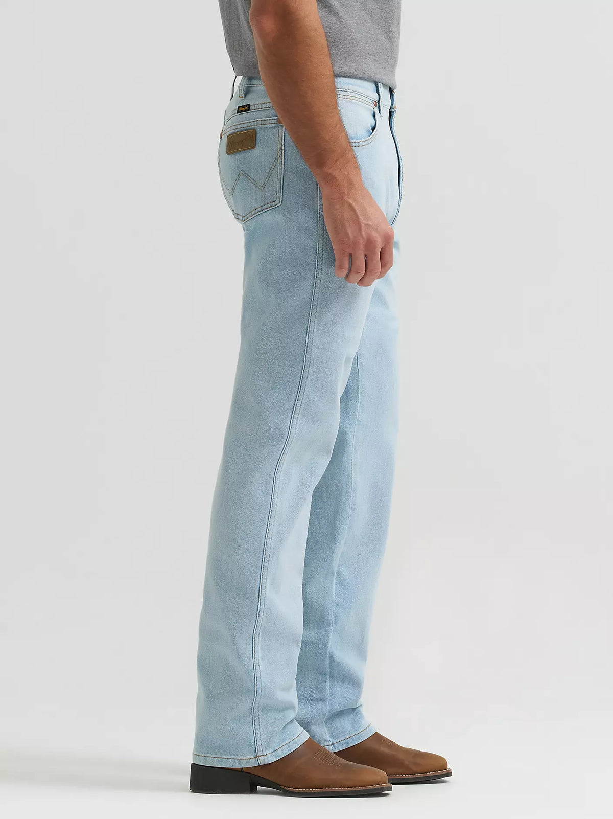 A side profile view of a person wearing light-wash blue jeans, a gray t-shirt, and brown leather shoes against a plain light gray studio background.