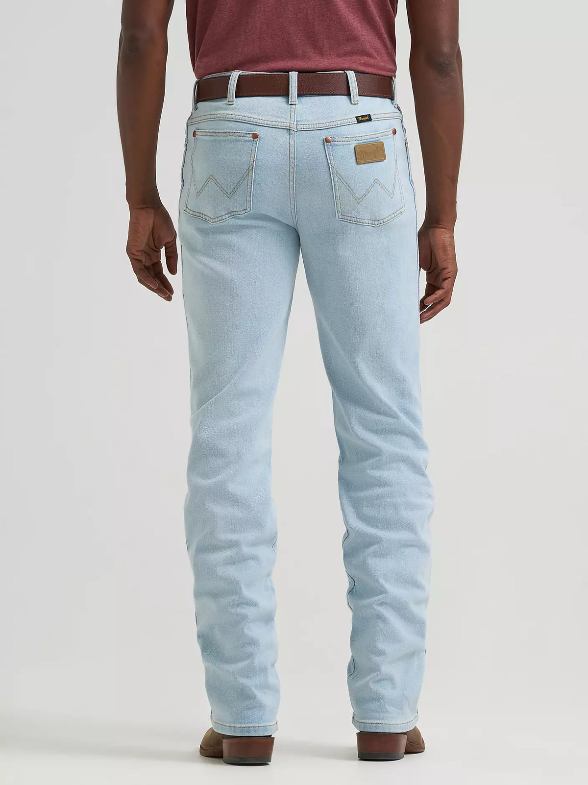 Rear view of a person wearing light-wash blue jeans with a brown belt and brown shoes, standing against a plain light gray studio background.