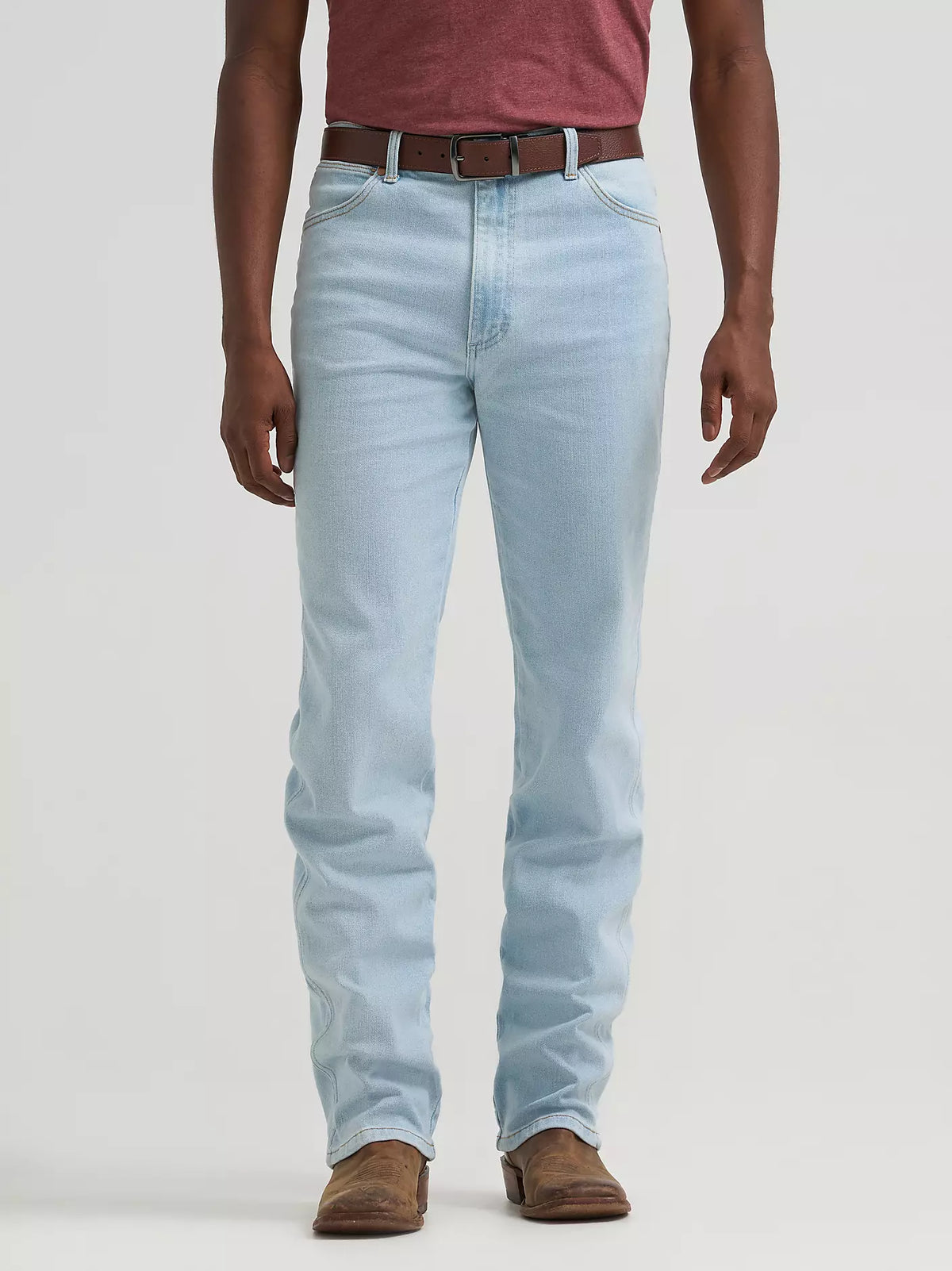 A person wearing light-wash blue jeans with a brown belt and brown shoes, shown from the waist down against a plain light gray studio background.