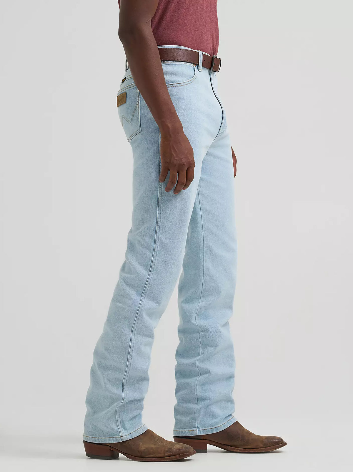 A side profile view of a person wearing light-wash blue jeans, a brown leather belt, a maroon shirt, and brown suede boots against a light gray studio background.