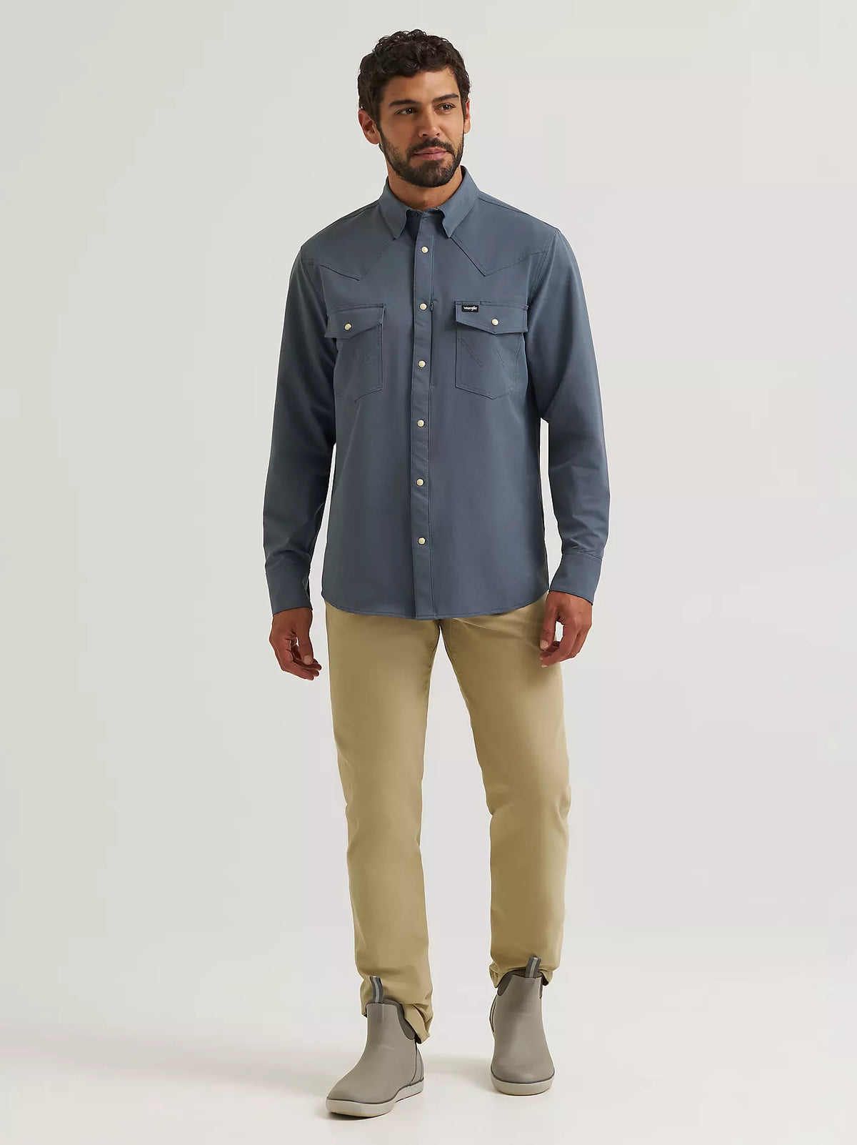 A man modeling a slate blue long-sleeve button-up shirt with chest flap pockets, paired with khaki pants and gray ankle boots, against a light gray studio background.