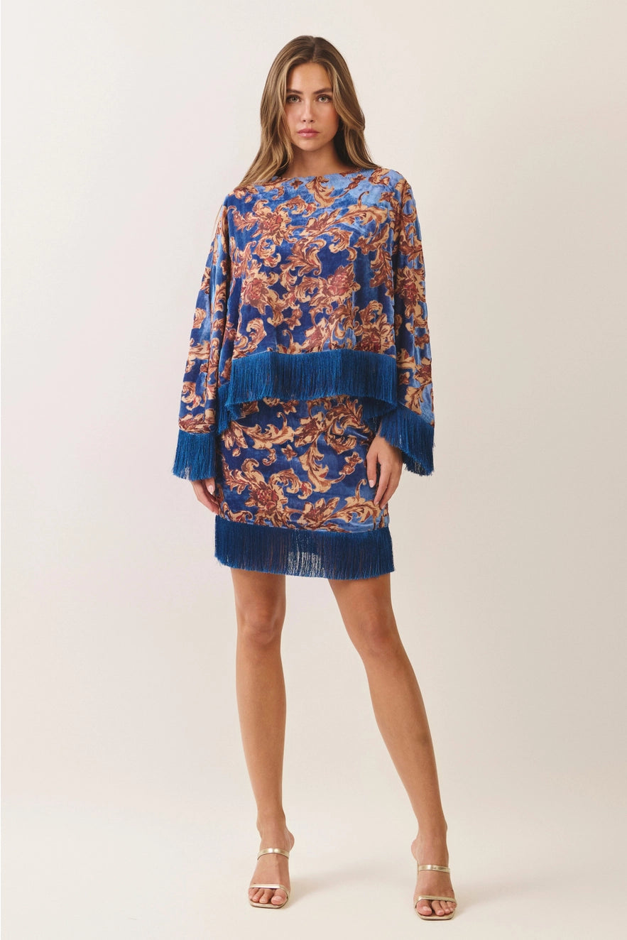 A woman modeling a blue long-sleeve mini dress with an orange and gold floral pattern and fringed hem and cuffs, photographed against a plain light beige studio background.