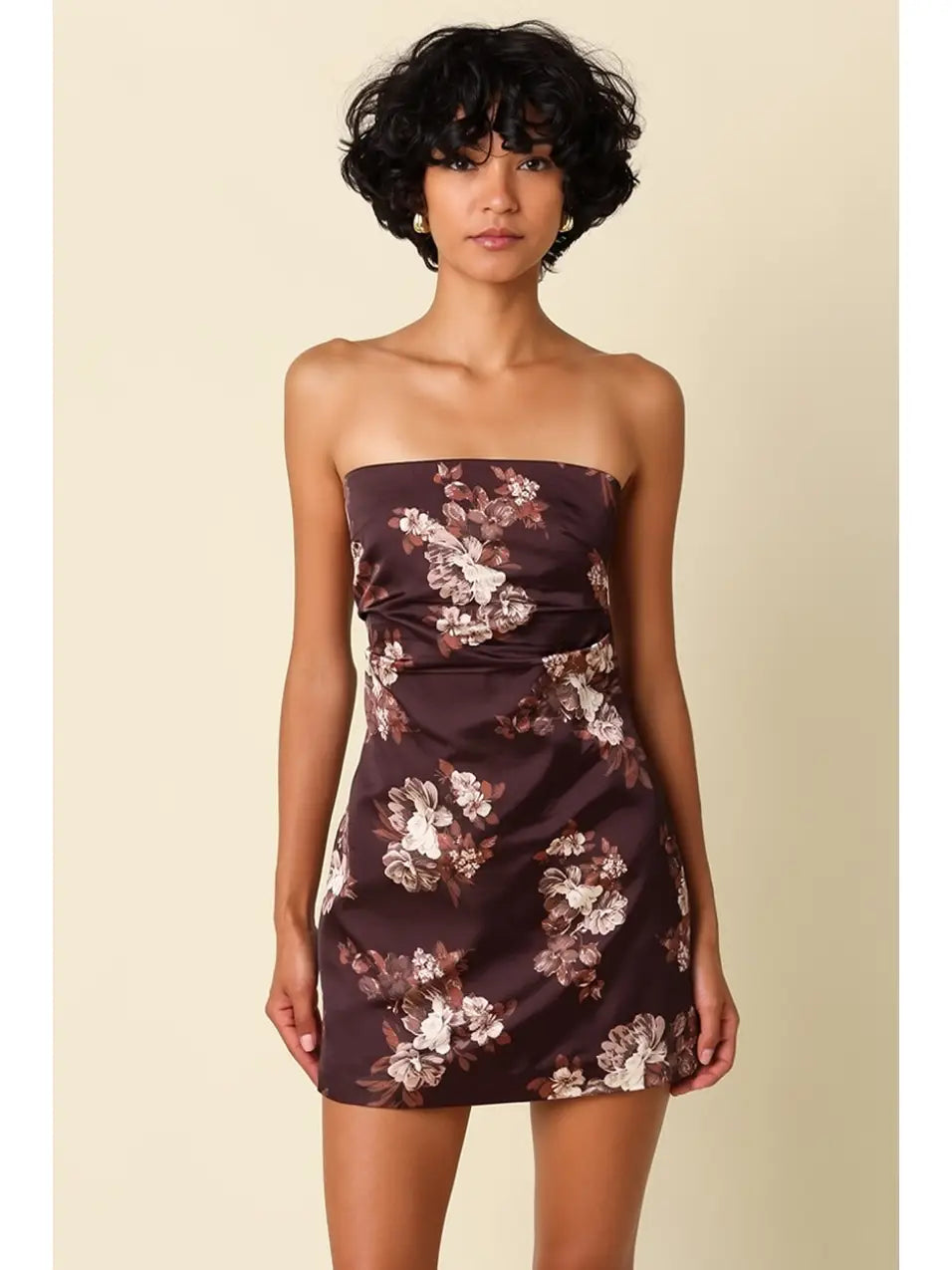A woman modeling a strapless mini dress with a dark brown base and a white and pink floral pattern, set against a solid beige background.