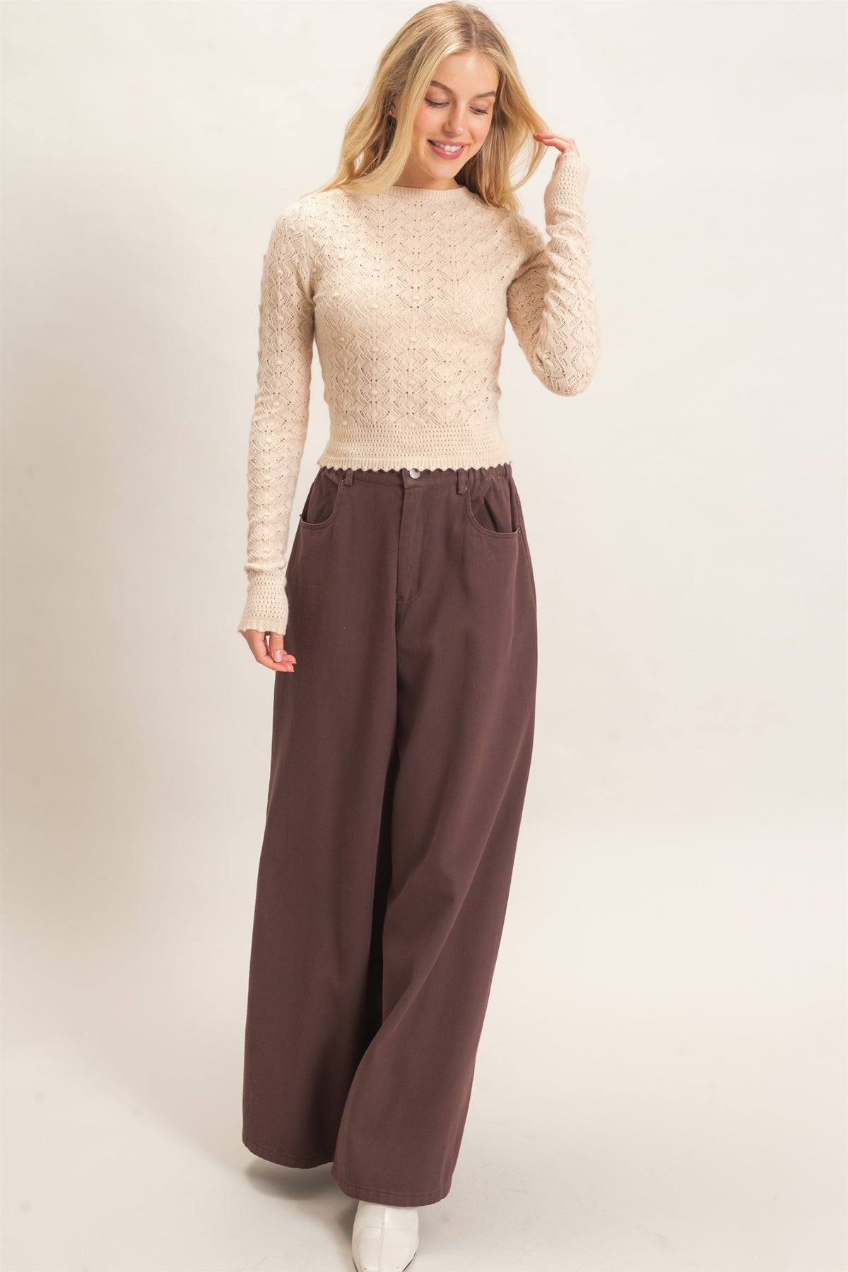 A woman modeling a cream-colored lace long-sleeve top paired with wide-leg brown trousers, standing against a plain light beige studio background.