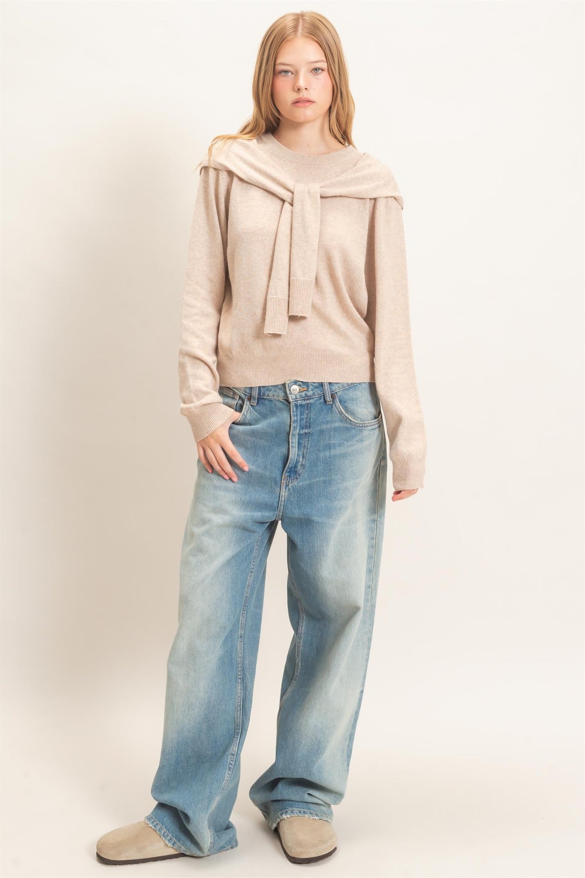 woman standing on white backdrop wearing beige sweater and jeans 