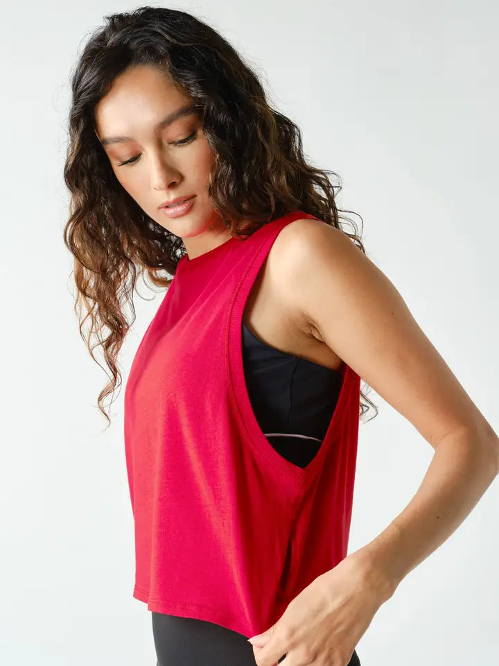 A woman wearing a bright red sleeveless crop top with a side cutout, layered over a black sports bra and black leggings, photographed against a clean white studio background.