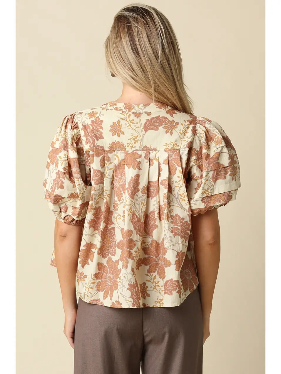 A woman viewed from the back wearing a cream-colored blouse with a dusty rose and brown floral pattern, featuring short puff sleeves and a gathered back. She is paired with brown trousers against a solid beige background.