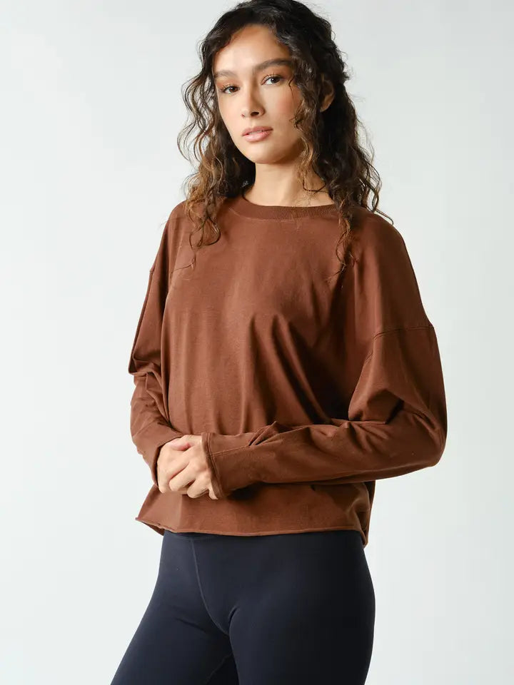 A woman wearing a brown long-sleeve crewneck top and dark navy leggings, standing against a light gray studio background.