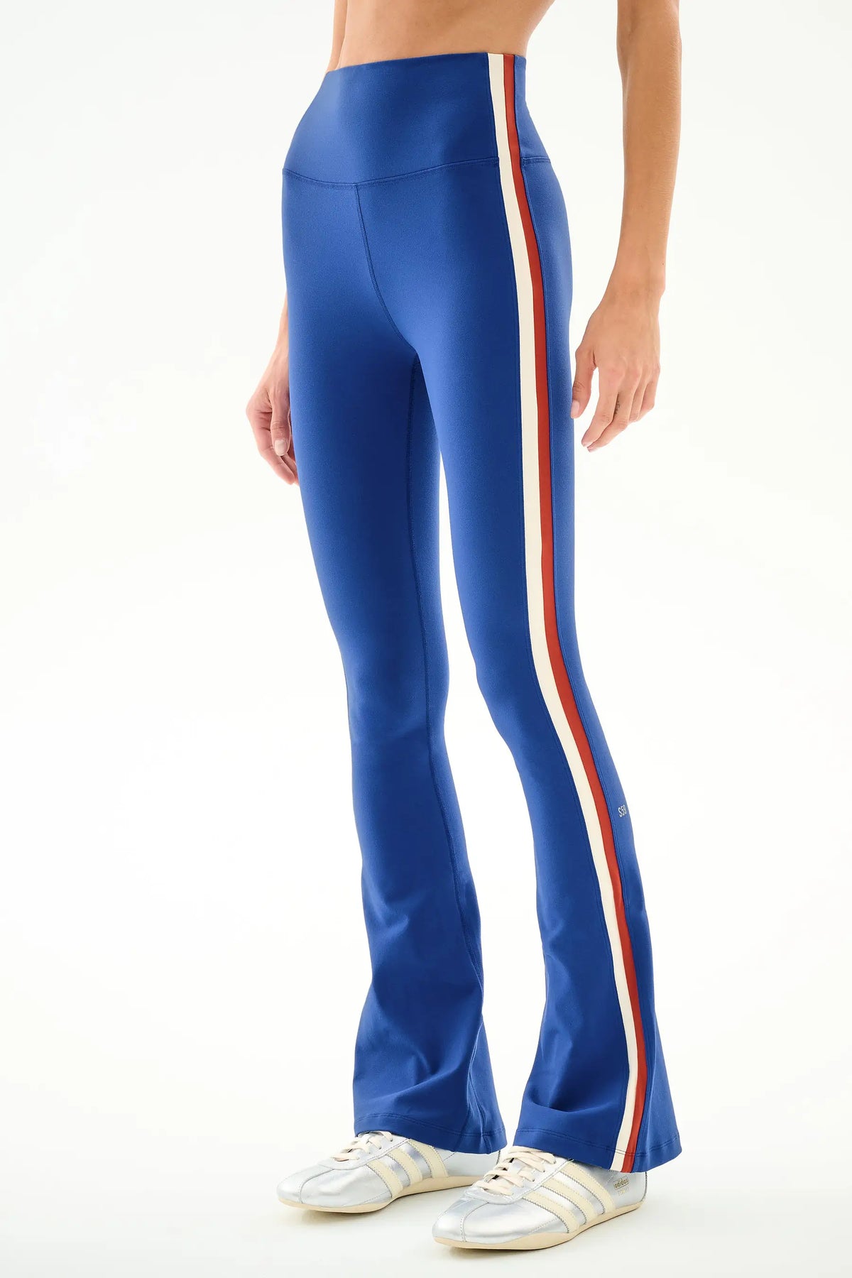 A model wearing high-waisted royal blue flared leggings with a vertical white and red stripe down the side, paired with white sneakers, against a plain white background.