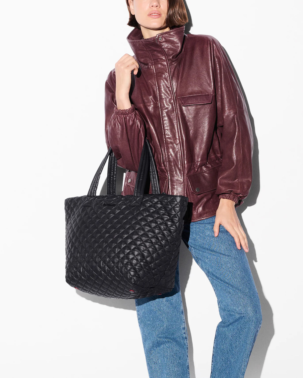 A model wearing a glossy burgundy faux-leather jacket and blue jeans carries a large black quilted tote bag against a plain white background.