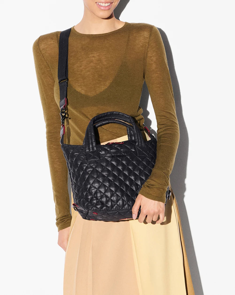 A woman wearing a brown long-sleeve top and beige skirt carries a black quilted top-handle handbag with a patterned strap against a white background.