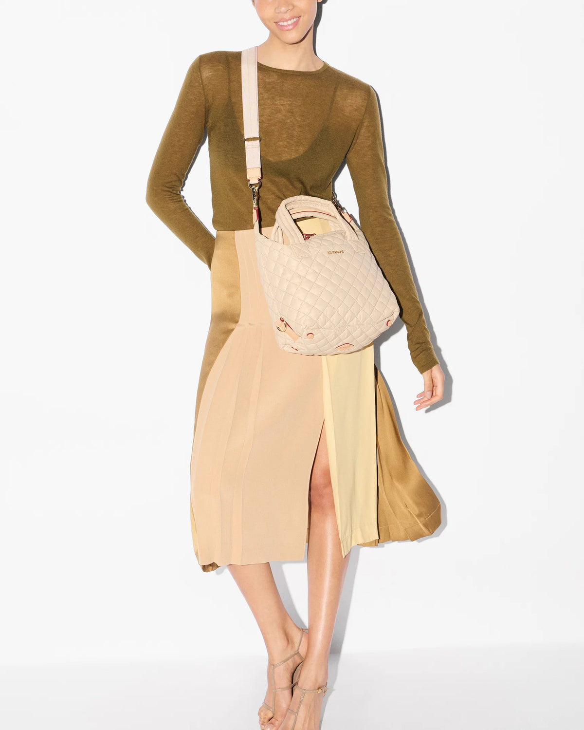A model wearing a brown long-sleeve knit top and a beige midi skirt with a side slit, carrying a quilted cream shoulder bag.