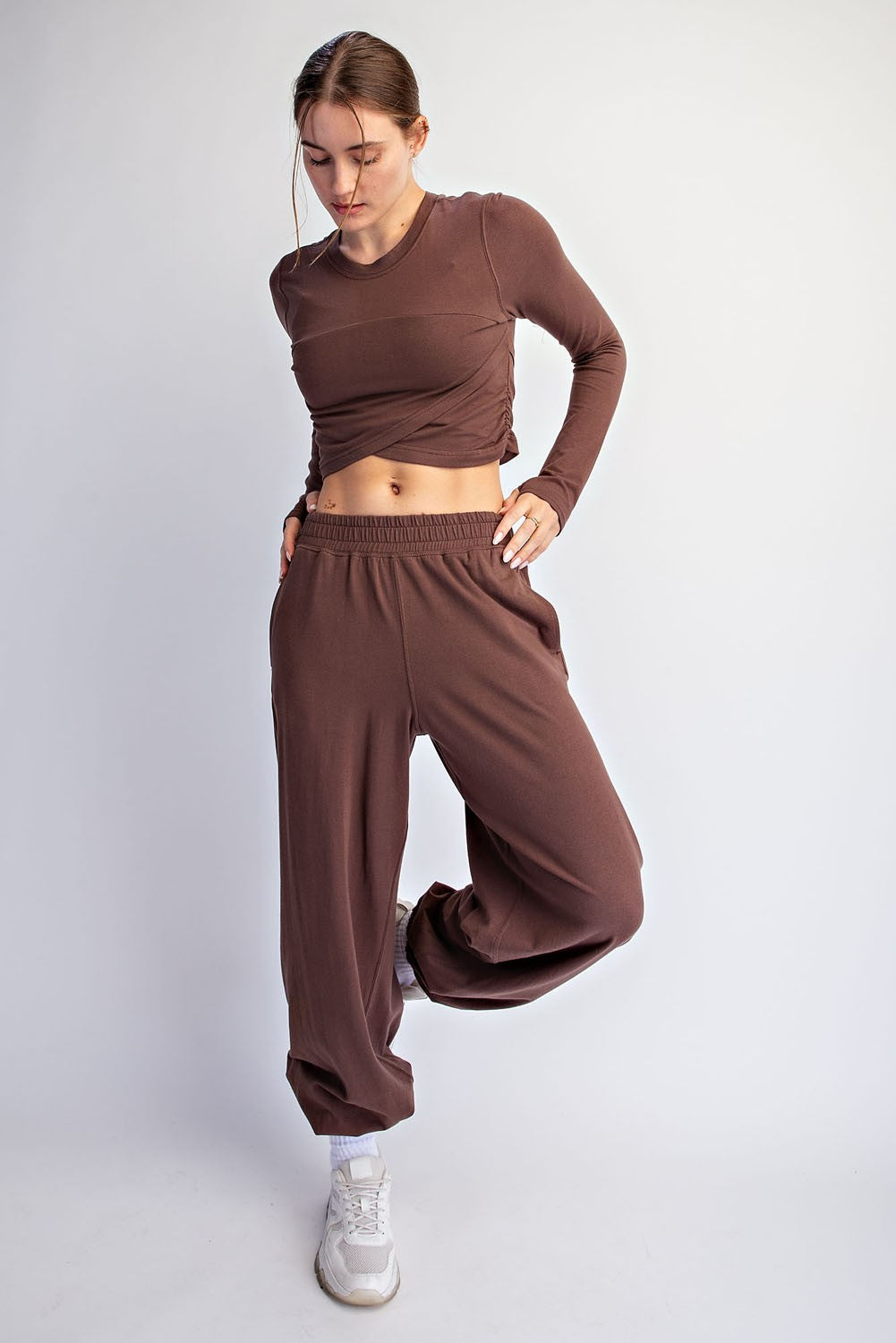A woman modeling a matching brown activewear set consisting of a long-sleeve cropped top and wide-leg sweatpants, paired with white sneakers, against a plain light gray studio background.