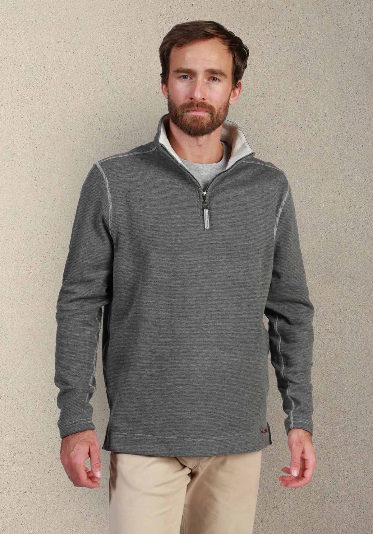 A man wearing a dark gray quarter-zip fleece pullover with a light gray inner collar, paired with beige pants, standing against a textured light gray wall.