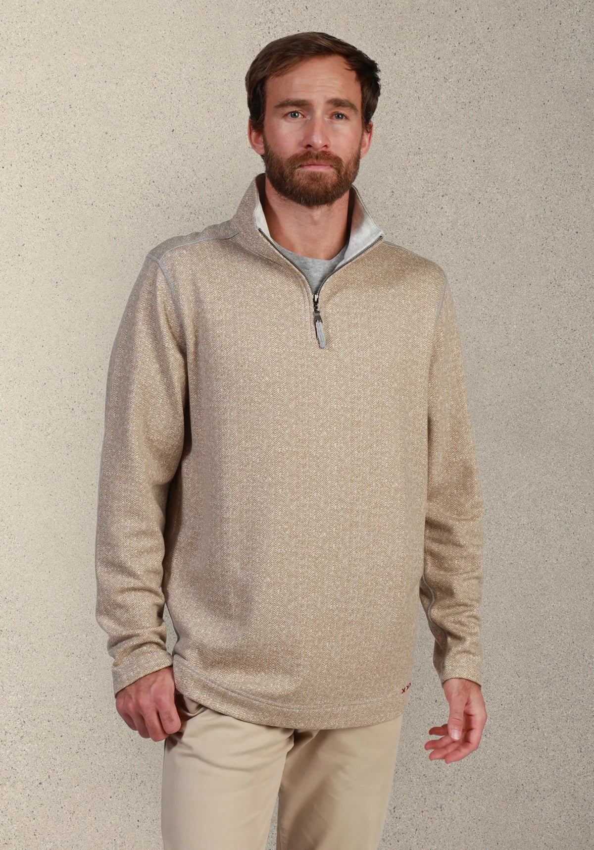 A man wearing a light beige heathered quarter-zip pullover sweater with a gray inner collar, paired with khaki pants, standing against a textured beige wall.