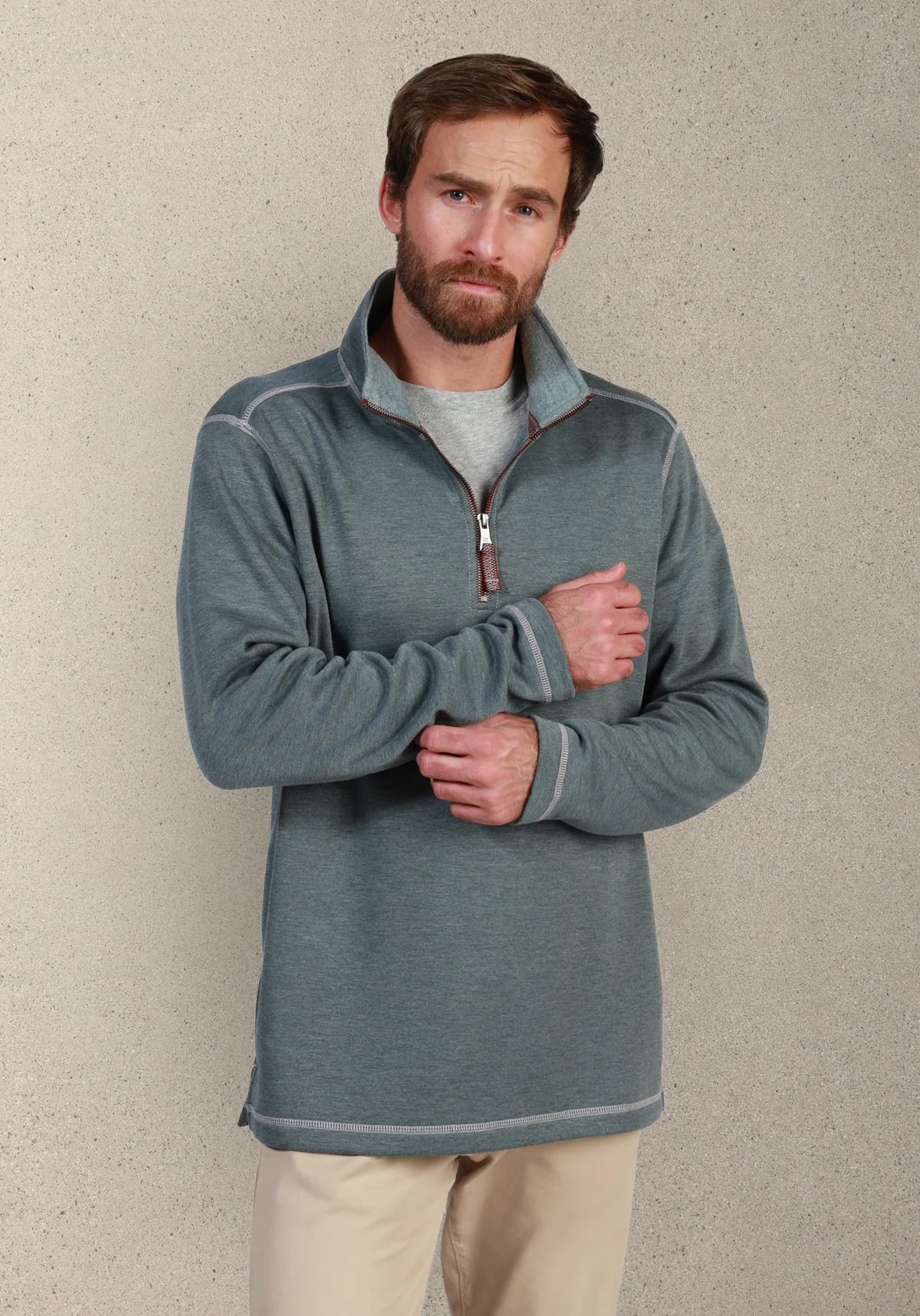 A man wearing a heathered slate blue quarter-zip fleece pullover sweater over a gray t-shirt and beige pants, standing against a textured beige wall.