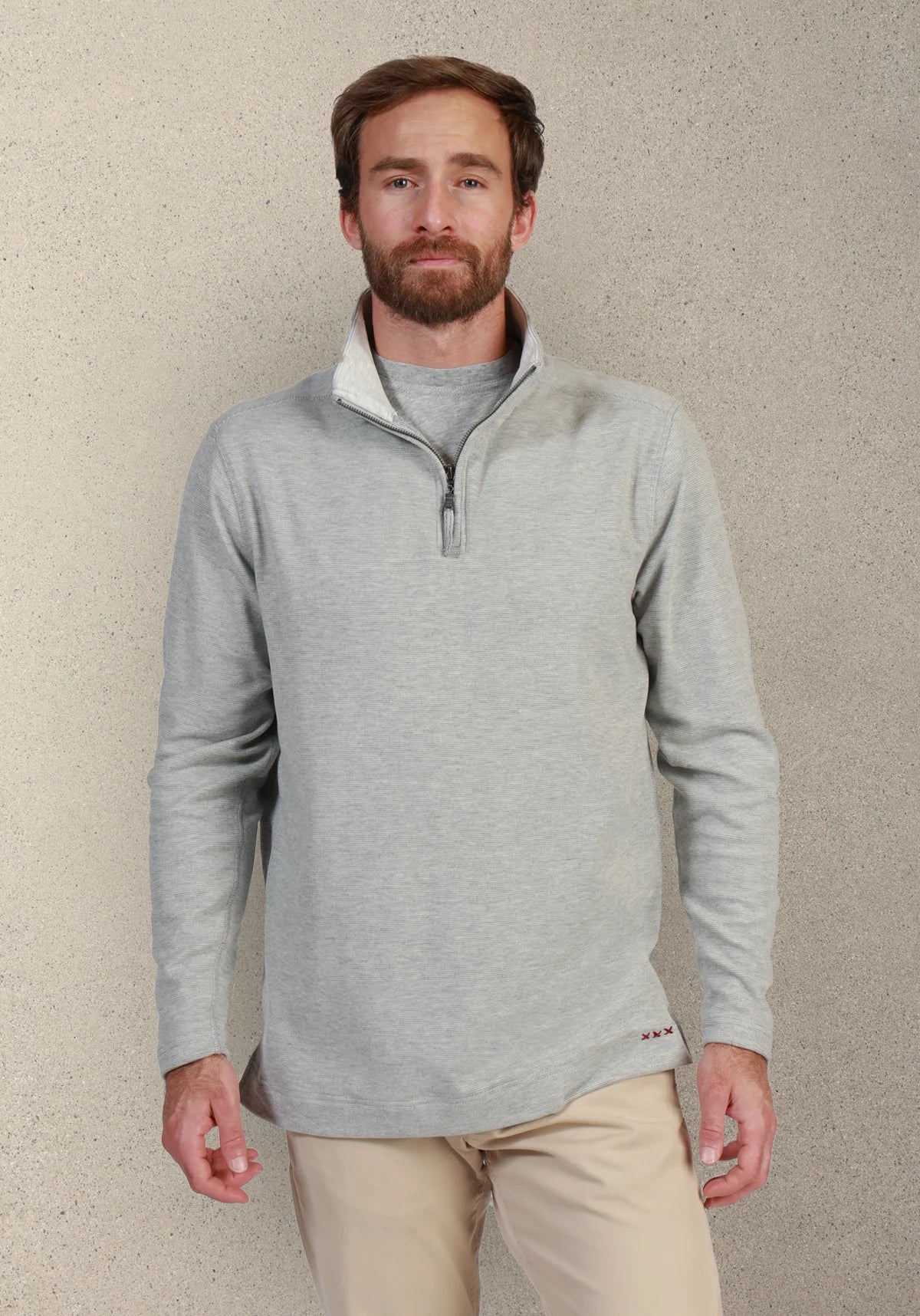 A man wearing a light gray quarter-zip pullover sweater with a white inner collar, paired with beige pants, standing against a textured beige wall.
