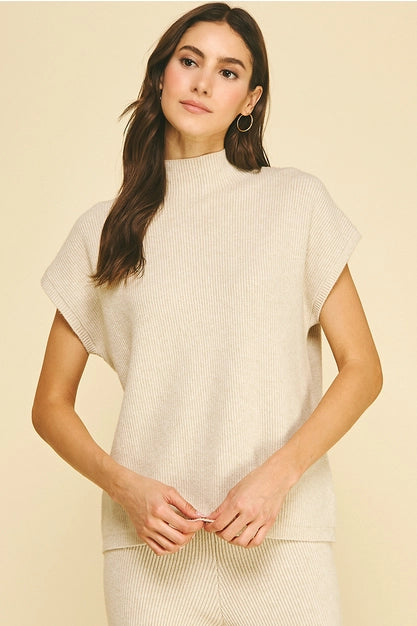 A woman modeling a ribbed knit two-piece lounge set in oatmeal, featuring a short-sleeve mock neck top and matching pants, set against a solid beige background.