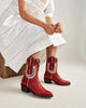 A person wearing a white dress sits on a wooden stool, showing off a pair of red western-style ankle boots with white embroidered details. The photo is taken in a clean, neutral studio setting.