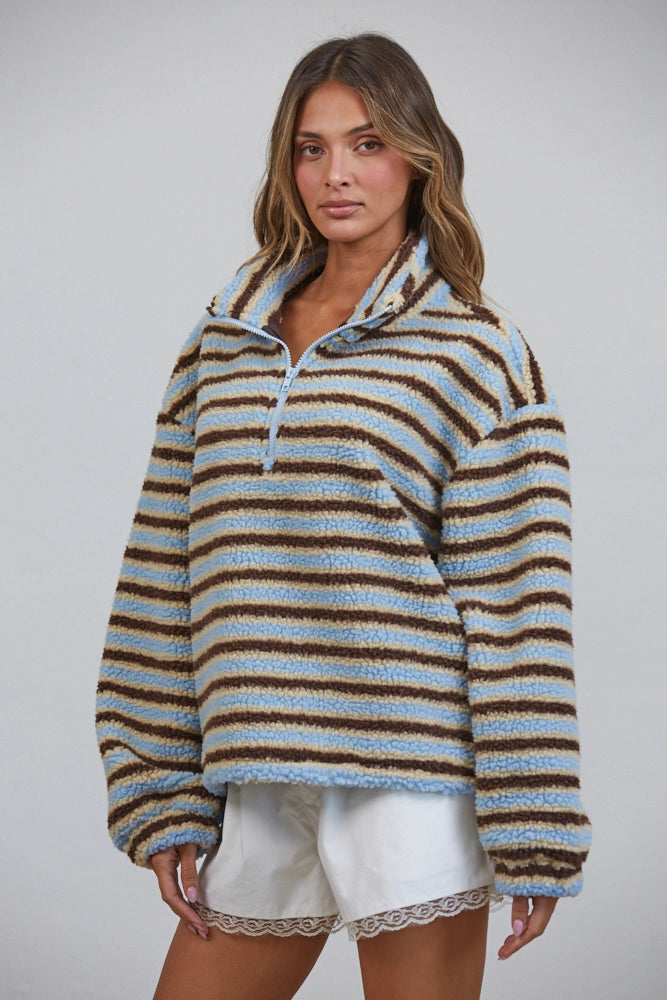 A woman modeling a fuzzy quarter-zip pullover sweater with horizontal brown, beige, and light blue stripes, paired with white shorts, against a solid light gray background.