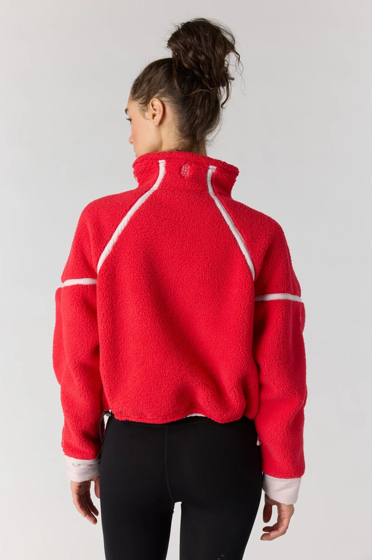 A woman viewed from the back wearing a bright red cropped fleece pullover with white contrast piping and black leggings, against a light gray studio background.