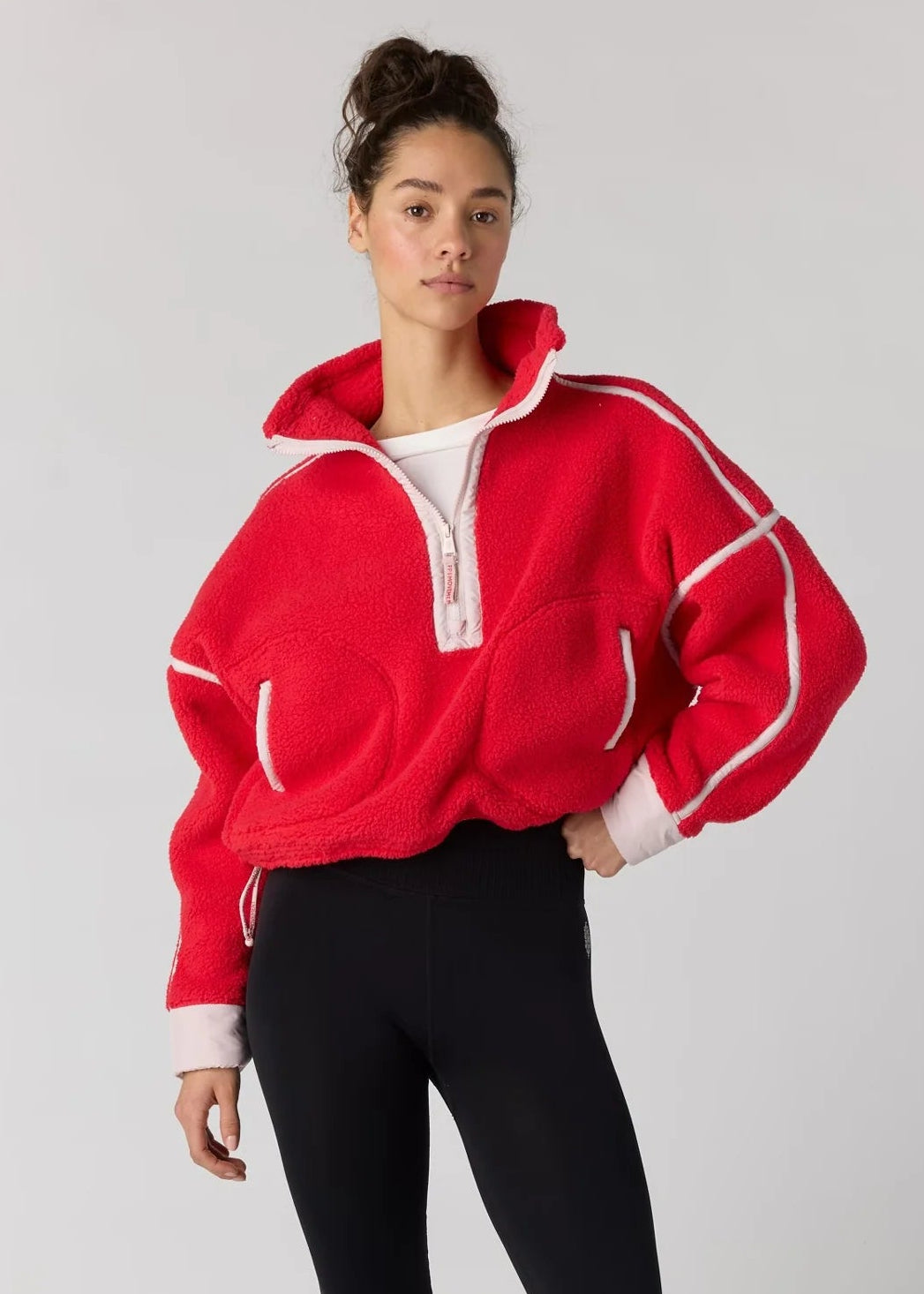 A woman modeling a bright red cropped quarter-zip sweatshirt with white contrast stitching and white ribbed trim, paired with black leggings and dark sneakers, against a light gray studio background.