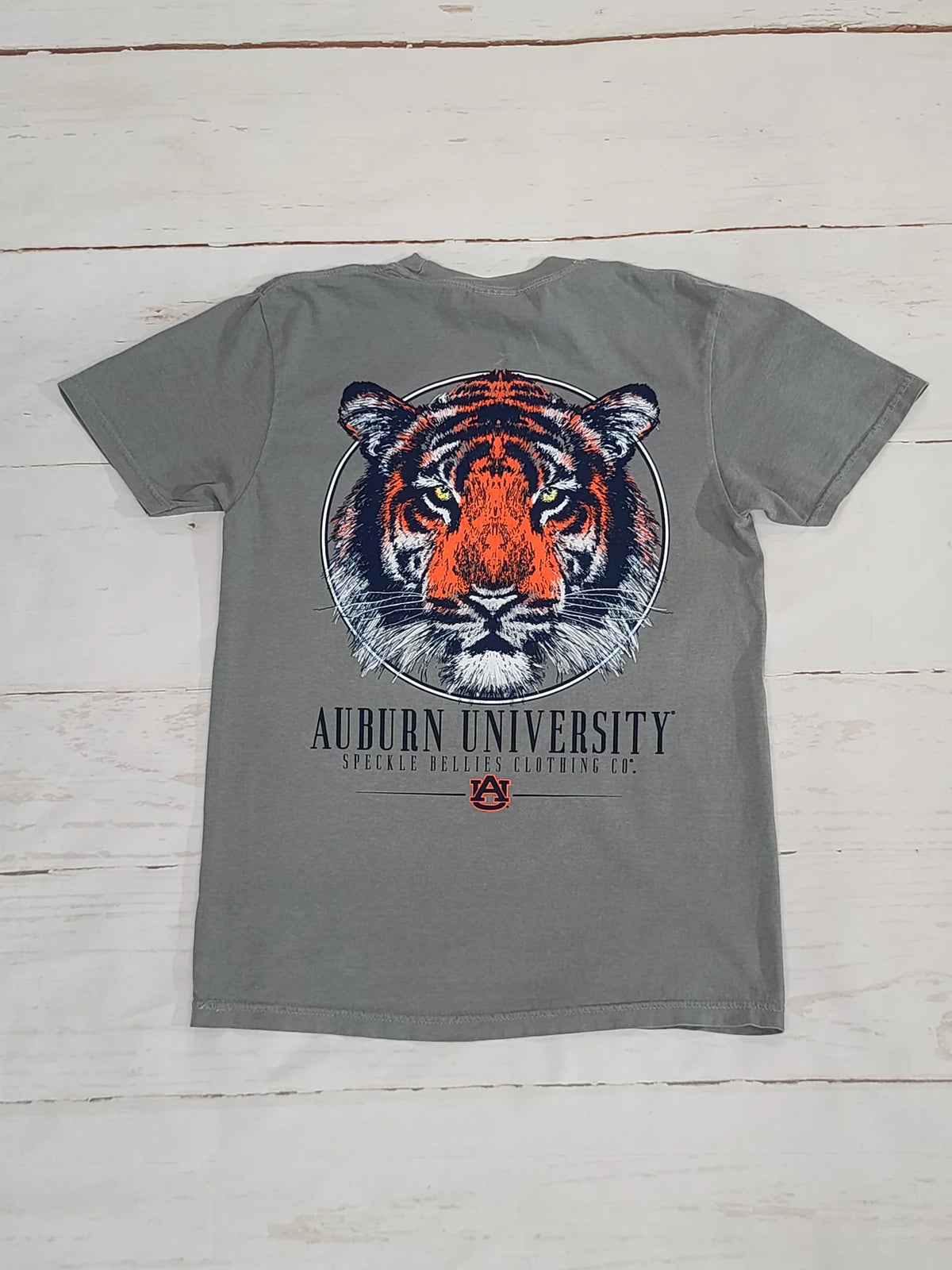 A gray short-sleeve t-shirt featuring a large graphic of an orange and black tiger face inside a circular frame, with 'AUBURN UNIVERSITY' printed below in black and a small Auburn logo. The shirt is laid flat on a white wood plank background.
