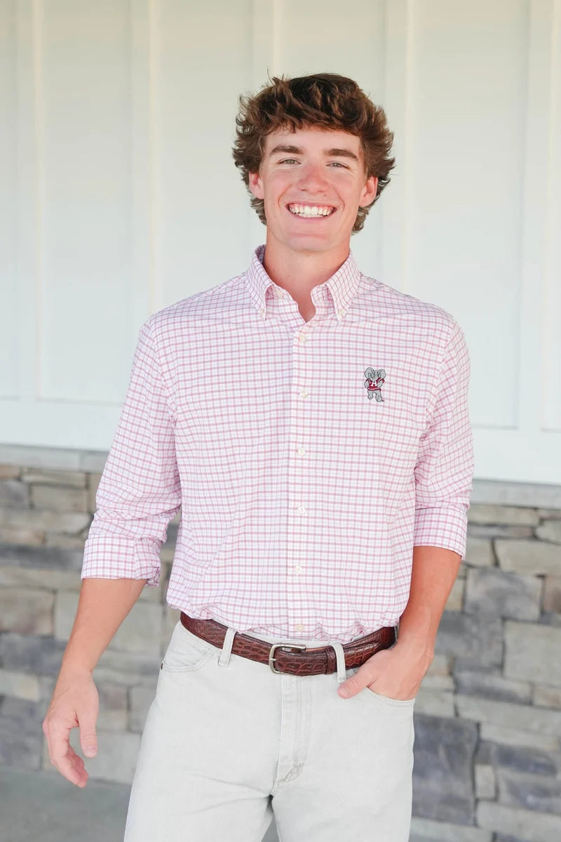 A smiling young man wearing a pink and white gingham button-down shirt with a small embroidered logo on the chest, paired with light beige pants and a brown belt, standing in front of a white paneled wall and stone base.
