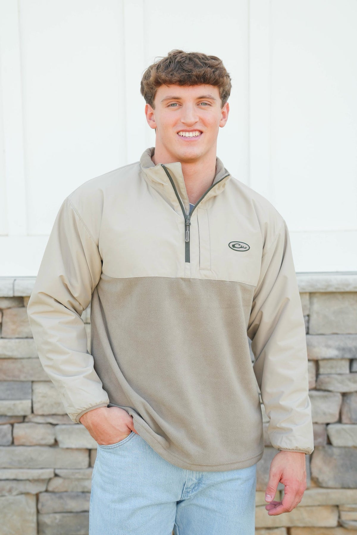 Man wearing a beige pullover with a logo, standing against a stone wall.
