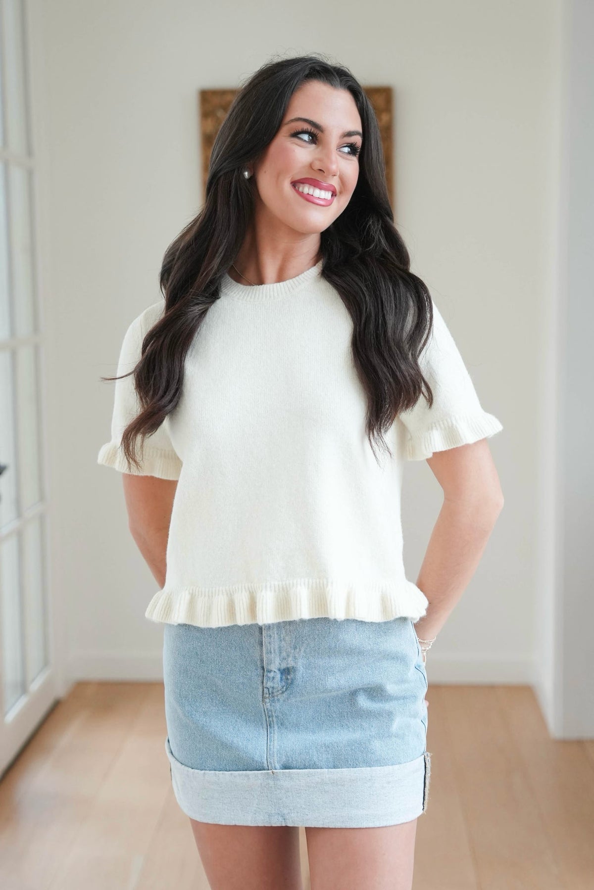 Woman wearing a white blouse with ruffled sleeves and a denim skirt indoors.