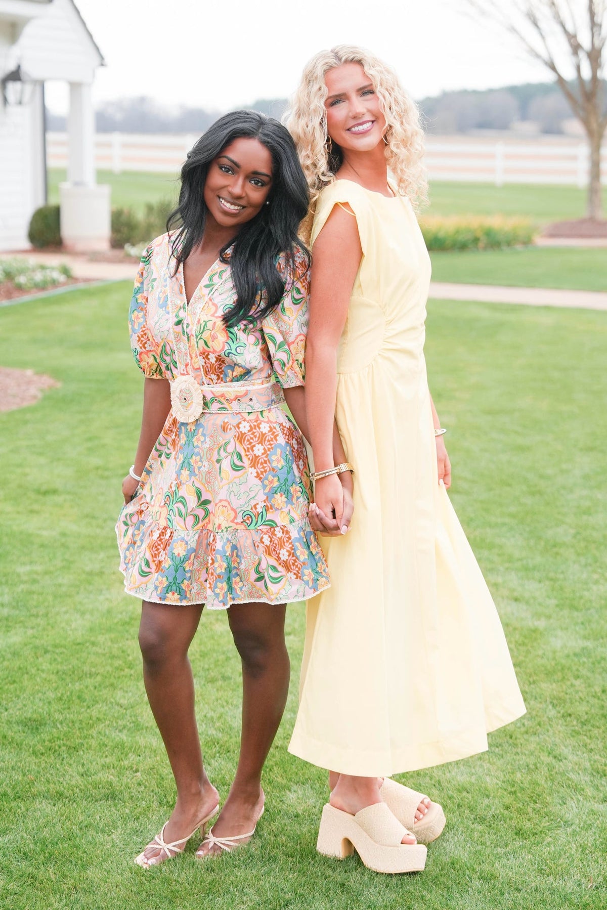 Two women standing on grass, one in a floral dress and the other in a yellow dress.