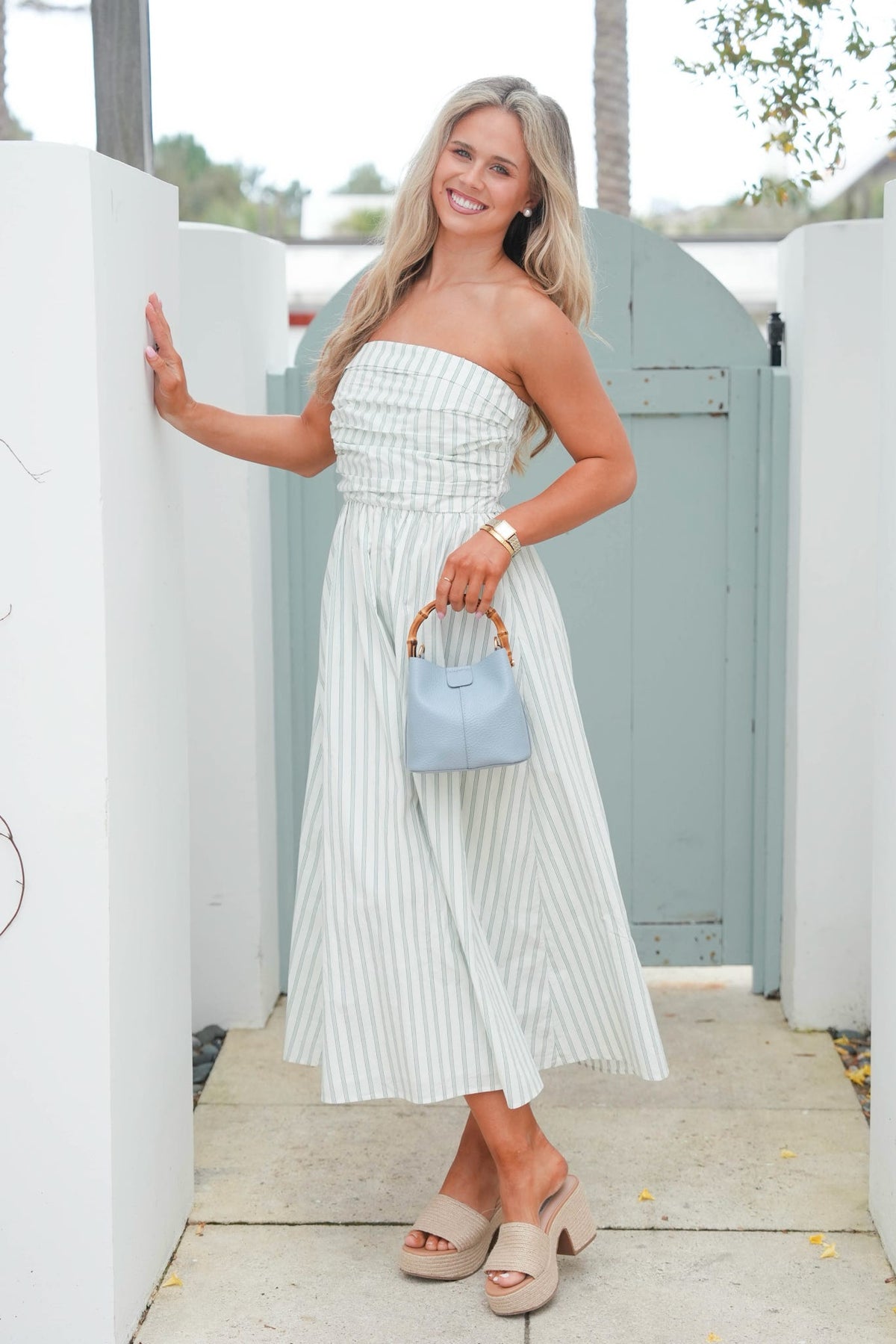 Woman in a strapless white dress holding a blue handbag, standing in front of a white wall and green door.